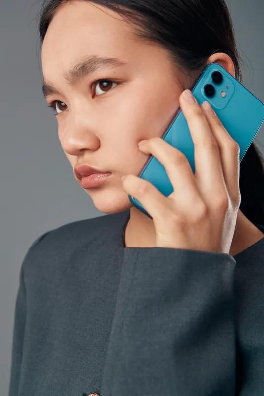 Young woman holding smartphone to ear for conversation or communication. Closeup portrait with serious expression and casual clothing against light background.