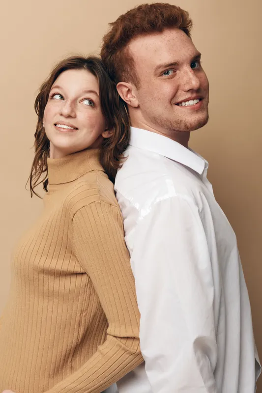 Smiling young man and woman stand back to back against beige background. Casual clothing and relaxed expressions create a friendly and warm atmosphere. Concept of friendship and connection.