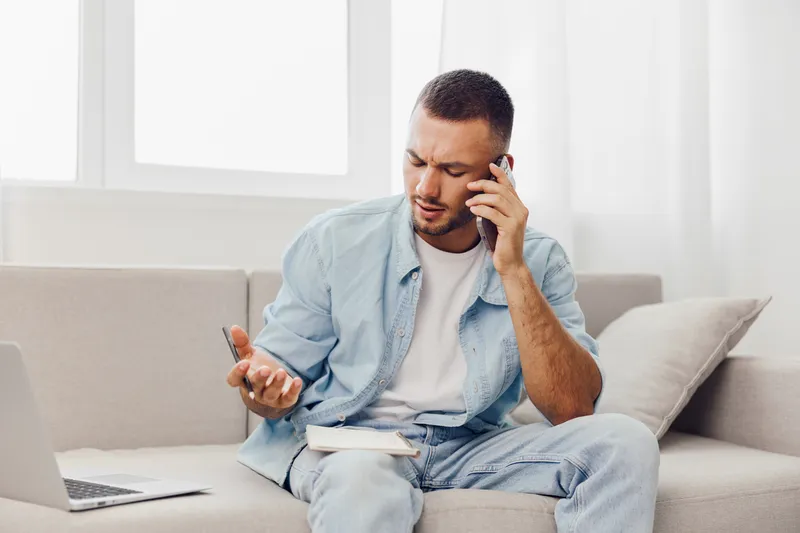 Thoughtful man on the phone, dressed casually in denim, sits on a comfortable couch with a laptop and notebook, showcasing modern work-from-home vibes and productivity.