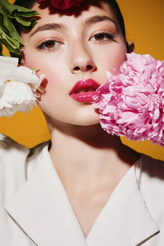 Close-up of a young woman with bold makeup and glossy lips surrounded by vibrant flowers on a warm orange background. Creative fashion beauty concept with hairy detail.