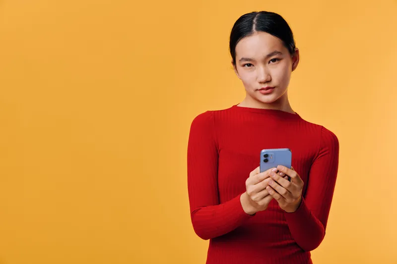 Young woman in red sweater holding smartphone, looking at camera with neutral expression on orange background. Female with dark hair using mobile device for communication or social media.