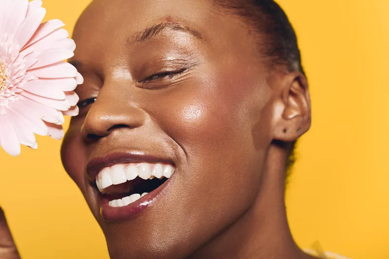 Close-up of happy African woman with dark skin smiling with closed eyes against yellow background holding pink flower. Concept of natural beauty, skincare, joy, and wellness.