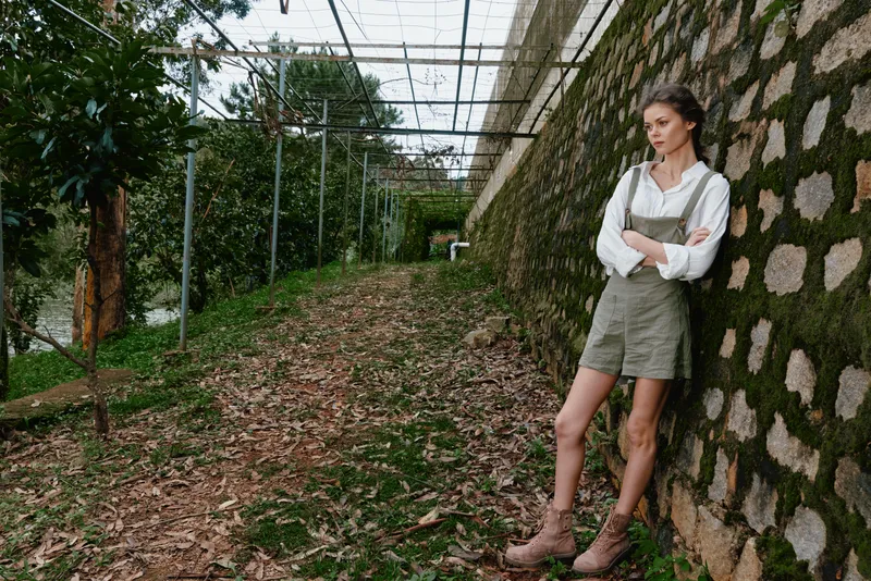 Young woman in casual outfit standing against a stone wall, showcasing a relaxed emotion in a lush outdoor setting, perfect for seasonal lifestyle concepts