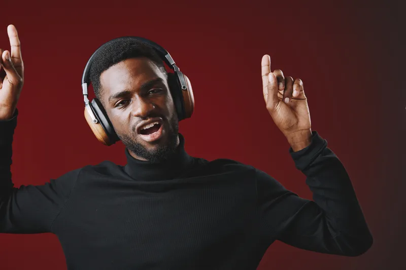 A joyful young African man in a black turtleneck wearing headphones, gesturing with enthusiasm, set against a warm red background, conveying happiness and energy