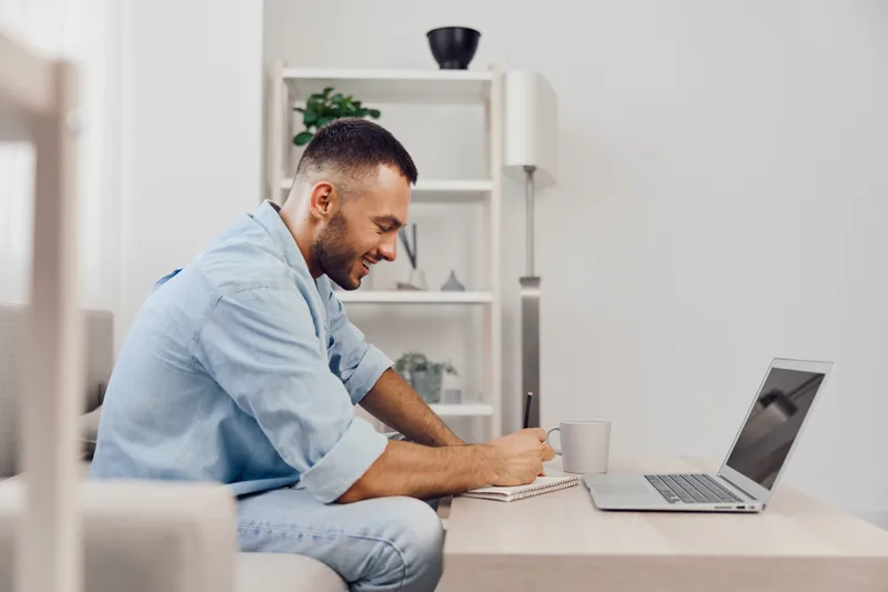 Smiling man working from home, sitting at a stylish desk with a laptop, creating a productive atmosphere filled with natural light and modern decor.