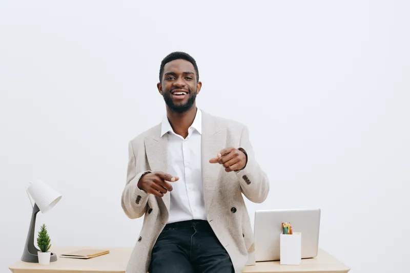 Smiling man in business casual attire, sitting at a modern office desk with a laptop, minimal decor, white background, relaxed and confident mood, suitable for corporate or professional concepts.