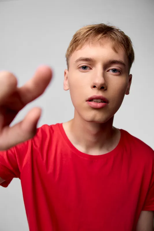 Close up of a young man reaching toward the camera in a red shirt, expressive face, dynamic gesture, casual studio shot with shallow depth of field and vivid lighting