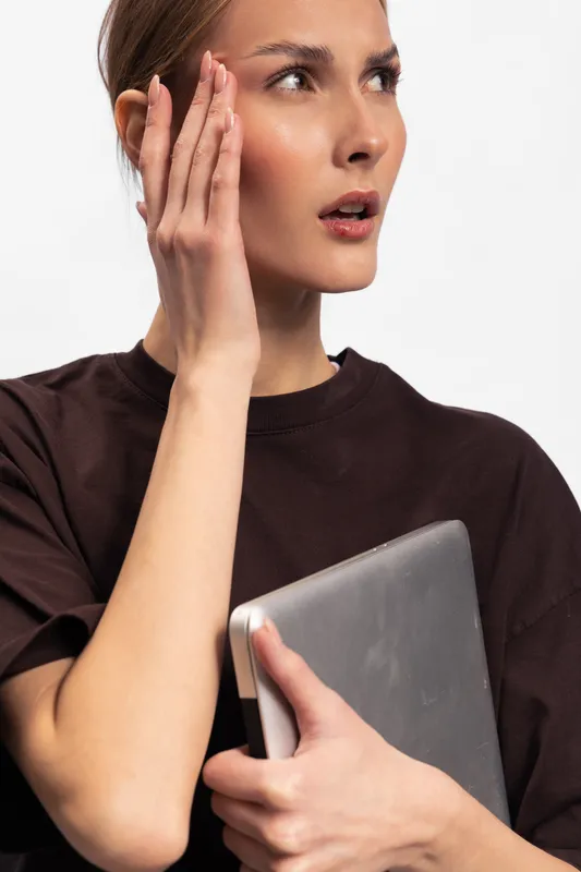 Young woman in dark shirt holds laptop and touches face with concerned expression. Isolated on white background. Concept of stress, anxiety, and digital work pressure.