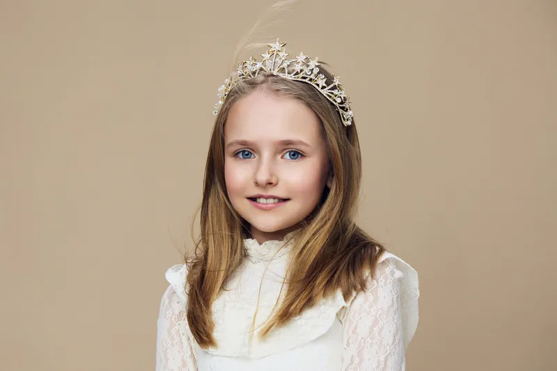 Charming young girl with a tiara and elegant dress, showcasing a joyful expression against a soft beige background, perfect for children s portraits and fashion themes
