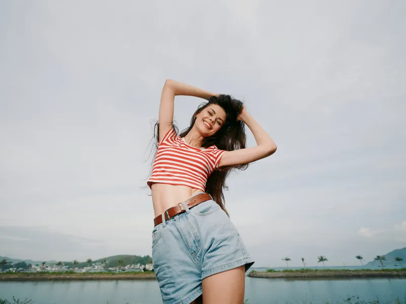 A young woman posing confidently outdoors, wearing a striped crop top and denim shorts, arms raised, sunlight bright, breeze in hair, capturing carefree summer energy.