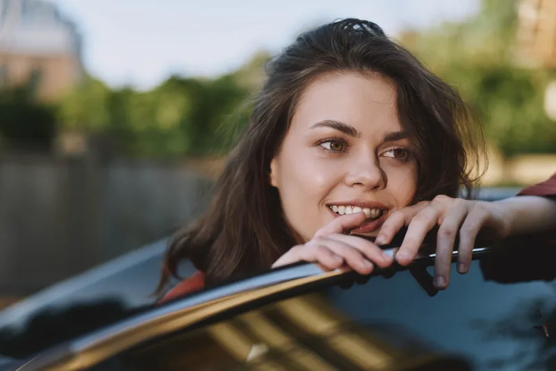 Young brunette woman rests her arms on a car window, smiling warmly under bright daylight in a casual outdoor setting, conveying relaxed everyday lifestyle and optimism