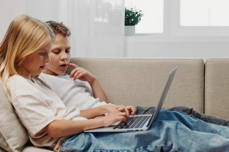 Two children sitting on a couch using a laptop together in a bright living room. Focused kids explore technology and digital learning in a comfortable home environment.