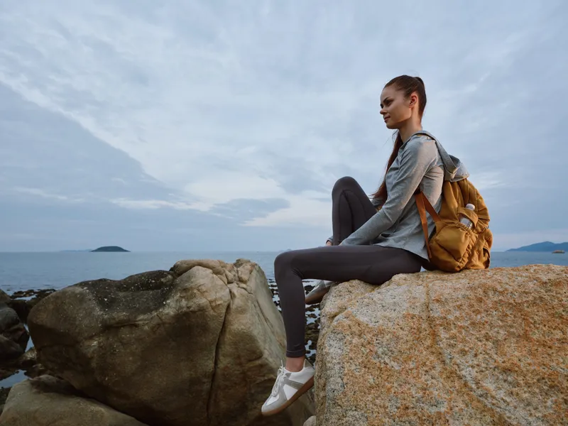 Adventurous woman in sporty attire sitting on rocks by the sea at dusk, showcasing a sense of tranquility and exploration with a backpack