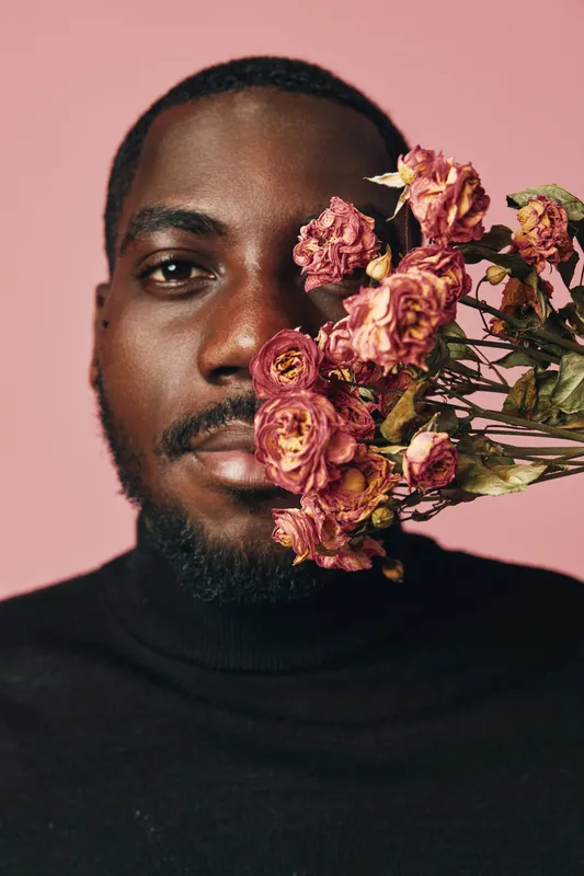 African man with beard and black turtleneck holding dried pink roses near face on pink background in creative conceptual visual style portrait with floral details and emotion.