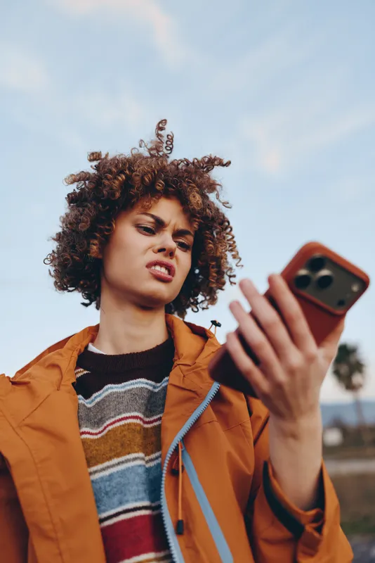 Woman selfie smartphone outdoors, curly hair and orange jacket, soft daylight sky background, casual lifestyle portrait, focused face holding phone and smiling slightly today