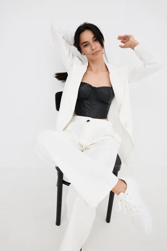 stylish woman in white suit and black corset sitting relaxed on chair against white background with casual sneakers