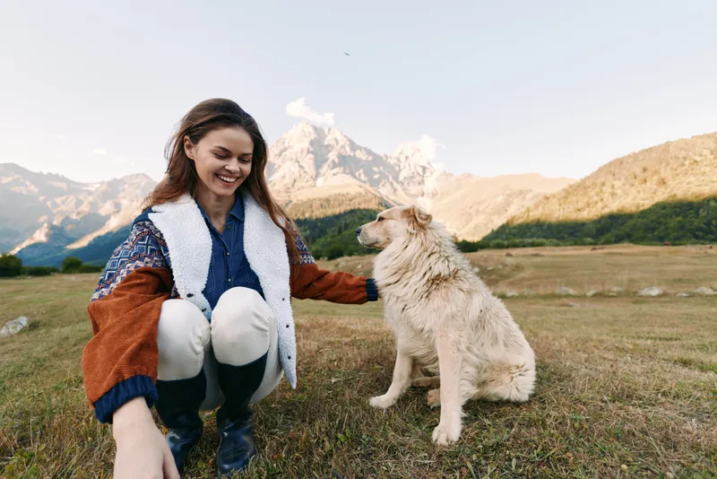Woman with dog in mountains and nature, pet interaction on outdoor meadow, smiling young traveler in warm jacket crouching and stroking friendly dog in scenic landscape.
