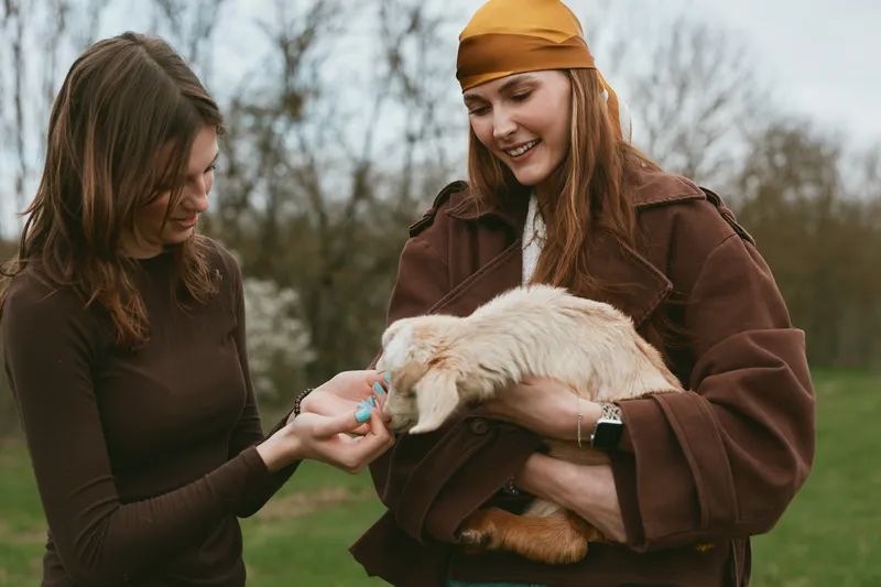 Two young women enjoy a playful moment outdoors as one feeds a baby goat held gently by the other in a natural spring setting with bare trees in the background.
