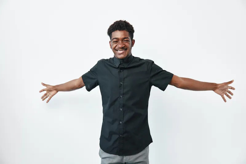 Young black man with dark skin smiling and standing with open arms in casual black shirt on white background for happiness and confidence.