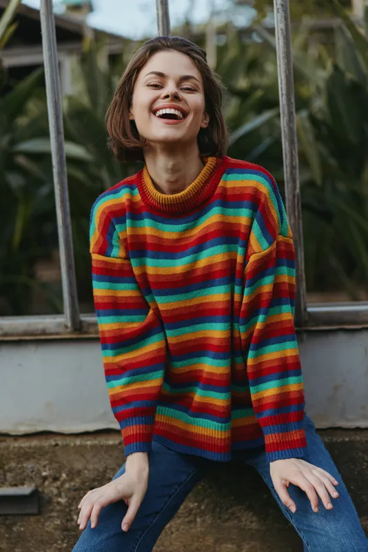 Woman smiling in a rainbow sweater portrait sitting outdoors in casual jeans. Young woman laughing at camera, vibrant knit jumper, relaxed pose, natural joy and carefree mood.
