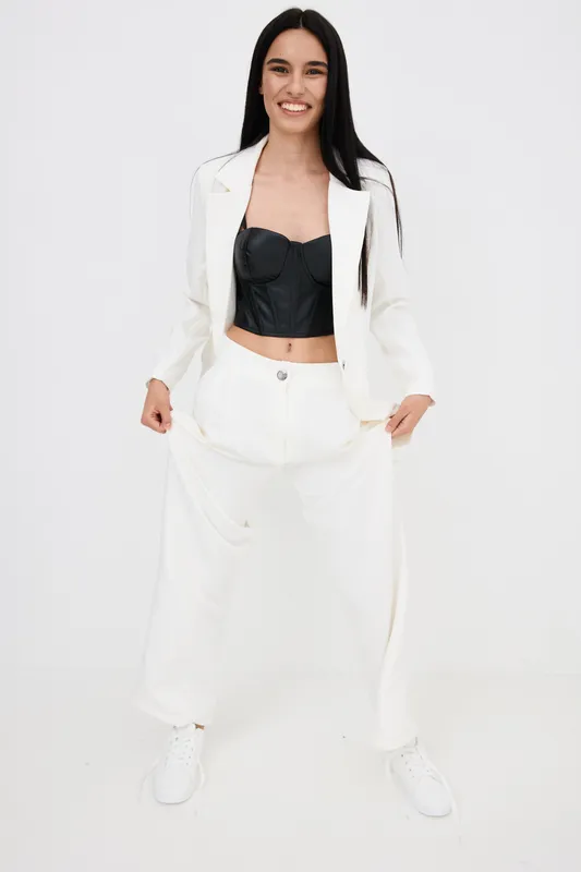 Smiling woman in white suit and black top standing confidently on white background, showcasing modern fashion with casual sneakers and relaxed pose.