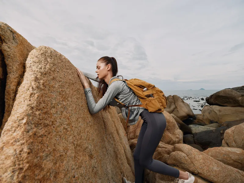 Young woman climbing rocks with a backpack, showcasing determination and adventure in an outdoor setting, perfect for travel and exploration themes