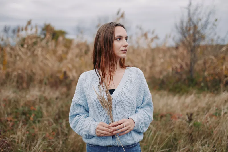Young woman stands in a windswept meadow holding dried grasses, wearing a knit sweater and jeans, a genuine portrait emphasizing natural, believable emotion and honest storytelling.