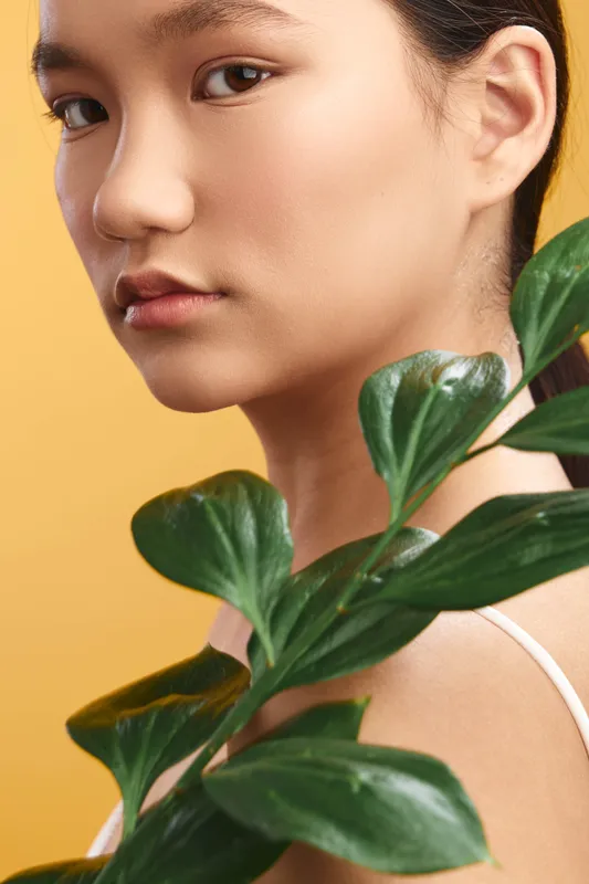 Asian woman gazes softly toward the camera amid lush green leaves with a warm yellow backdrop, presenting calm beauty, natural freshness, and a serene studio vibe.