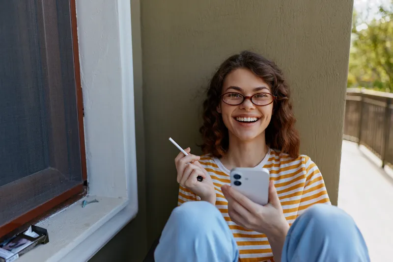 Cheerful woman indoors near a window, leaning against a wall, looking at her phone, wearing a striped shirt, relaxed through sunlight, contemporary home lifestyle moment, casual and bright