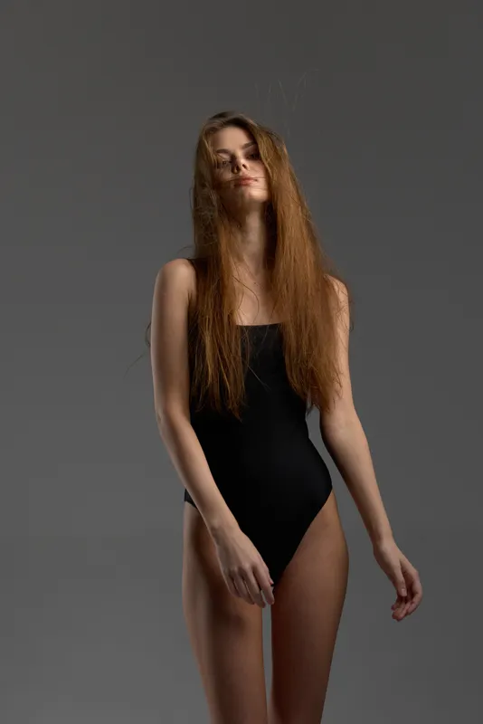 Young woman in black swimsuit posing with long hair against gray background, fashion, beauty, confident, studio lighting