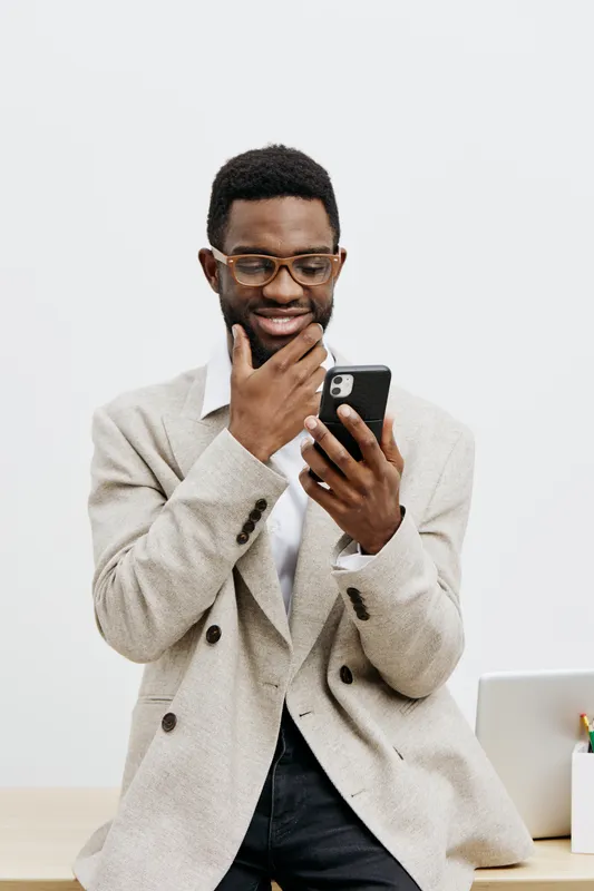 Smiling man using smartphone, dressed in beige blazer and glasses, in bright modern office with laptop on desk, casual business attire, cheerful expression, technology lifestyle concept, indoor settin