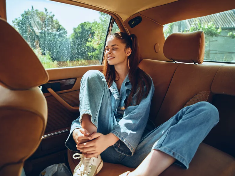 Young woman enjoying a joyful moment in the backseat of a car, wearing denim and smiling, with a bright green landscape outside the window.