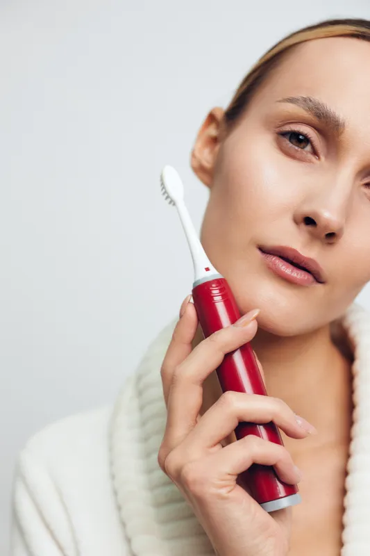 Woman in a plush robe holds a red electric toothbrush near her face, showcasing skincare and grooming in a bright clean setting, calm portrait with soft lighting