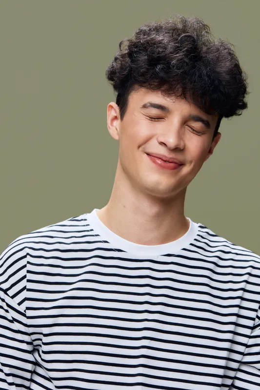 Smiling man with curly hair wearing a striped shirt, showing relaxed and happy expression against a soft olive green background, casual style portrait.