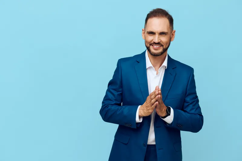 Professional businessman stands against a solid blue background, projecting confidence and calm as he joins hands in a thoughtful, composed gesture, ready for impactful communication.