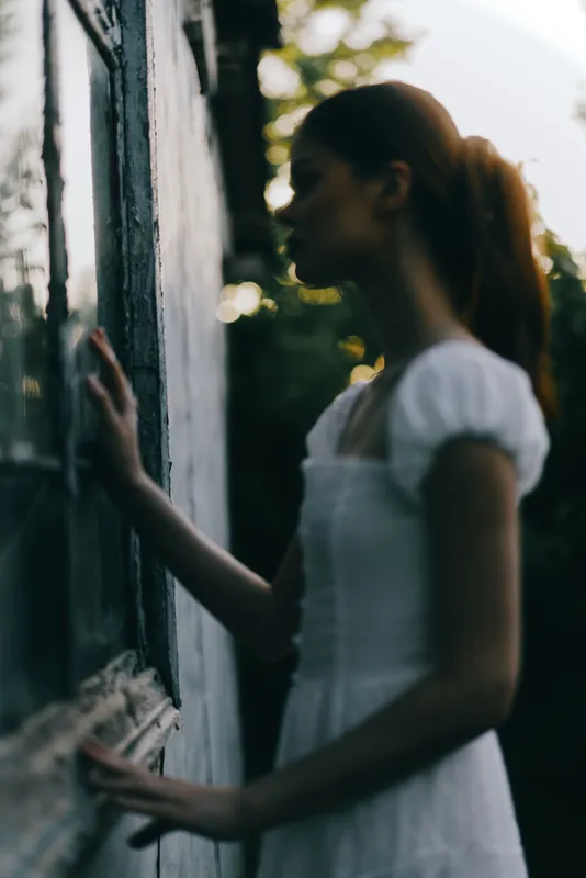 Young woman in a white dress gazing thoughtfully out of a window, capturing the essence of nostalgia and serenity in a natural setting