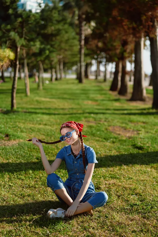 relaxed woman sitting on grass with braids wearing sunglasses and denim overalls in sunny park surrounded by trees and green lawn
