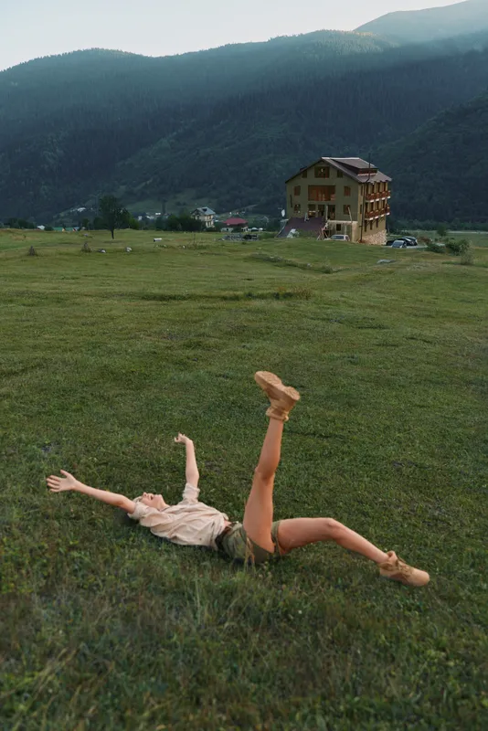 Field view shows a woman outdoors lying in grass, arms and legs cheerfully stretched, enjoying a carefree outdoor moment, rural hills and a house in the background, lifestyle candid