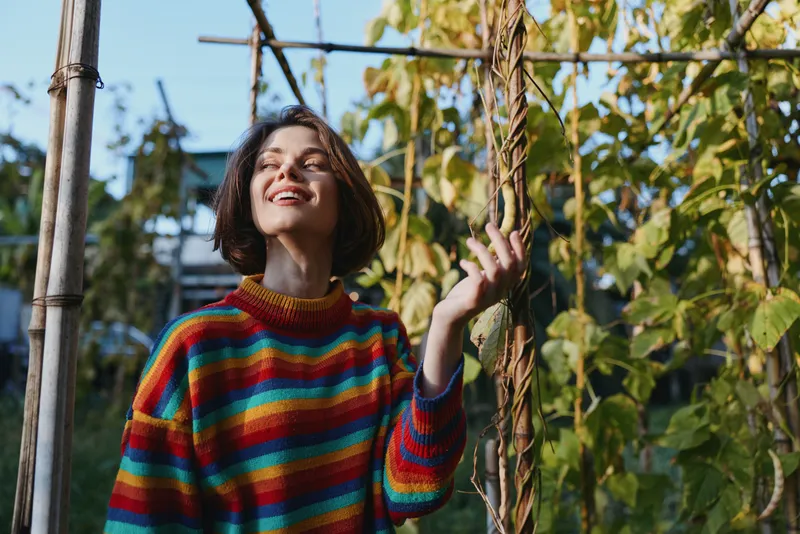 Woman in a colorful striped sweater smiling in a sunlit garden, young adult touching a vine on a wooden trellis, casual outfit and relaxed joyful expression for lifestyle and fashion.