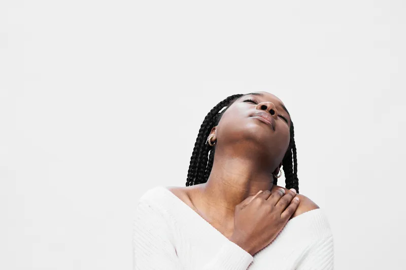 Young Black woman with braided hair expresses tranquility against a minimalist white background, wearing a cozy off shoulder sweater, highlighting serenity and calmness