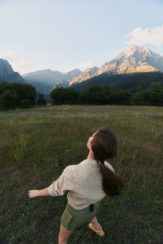 Young girl in light shirt and olive shorts runs through a grassy meadow with towering mountains in the background, conveying freedom and outdoor wonder.