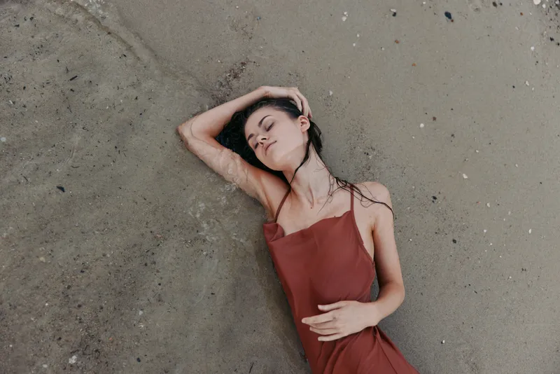 A peaceful portrait of a woman lying on soft sand by the shore in a rust red dress, eyes closed, relaxed pose, gentle light creating a warm, serene mood.