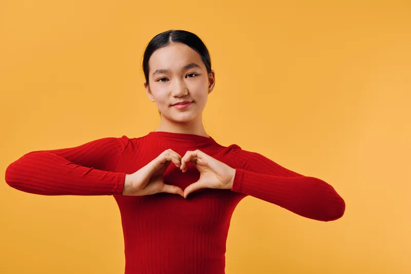 Young woman in red sweater making heart gesture with hands smiling at camera isolated on orange background. Portrait of female showing love and positive emotion with folded fingers.