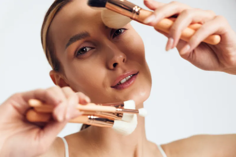 Close up of a woman applying makeup with brushes, showing makeup technique, soft lighting, focused expression, beauty routine in studio, candid mood natural skin tones and subtle glow.