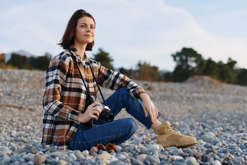 Woman with a camera sits on a pebbled beach, exploring the shoreline in casual denim and plaid shirt, capturing relaxed coastal scenery with boots, outdoorsy vibe and calm, scenic light.