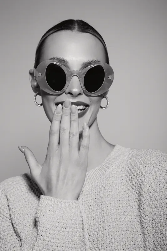 Young white woman with fair skin wearing round sunglasses and hoop earrings smiles, hand covering mouth in studio.