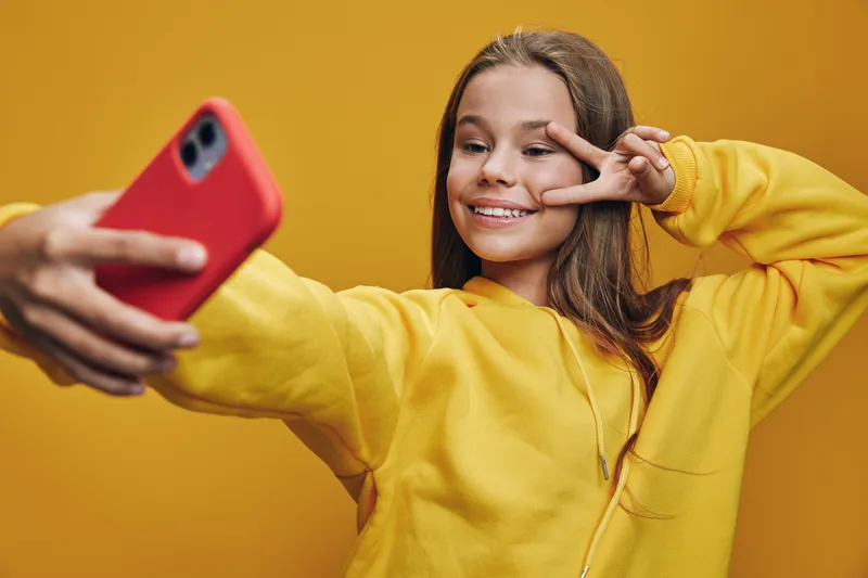 Girl in yellow hoodie taking selfie with smartphone making peace sign with fingers smiling on orange background cheerful lifestyle and youth culture.