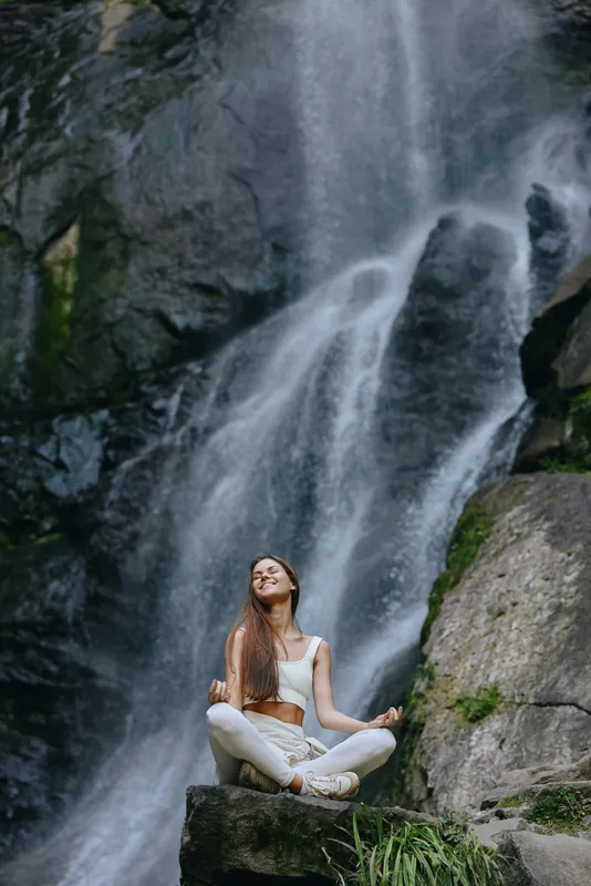 Young woman meditating beside waterfall in nature, wearing white clothes, sitting cross-legged on rock, serene and peaceful outdoor scene with lush greenery.