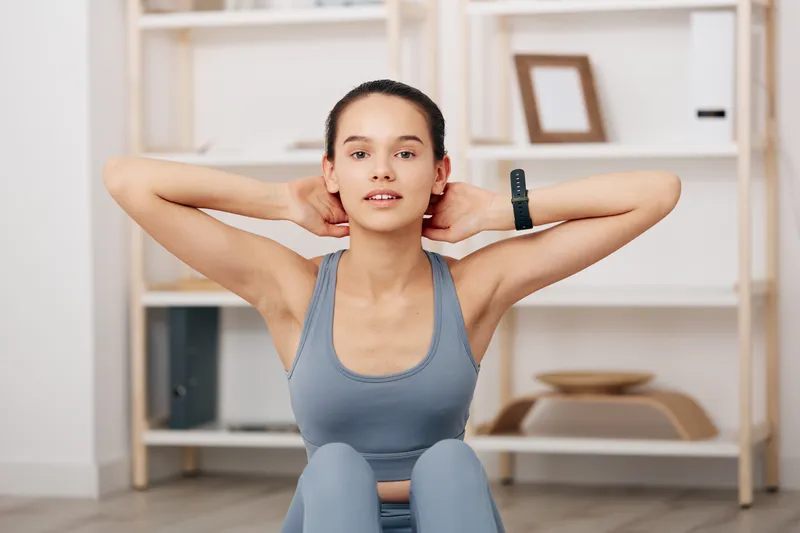 Woman in athletic wear performing sit-up at home, hands behind head, focused expression, mid core workout in a bright living room with shelves and minimal gym gear.
