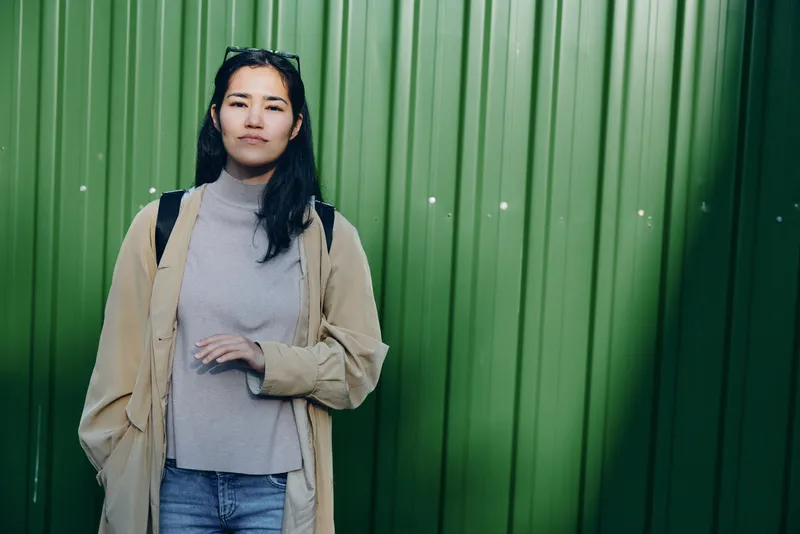 Casual woman standing against a vivid green wall, wearing a beige coat over a gray knit sweater and blue jeans, relaxed street style for everyday fashion and urban lifestyle imagery